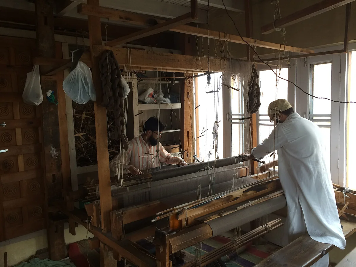 A photograph of a Kashmiri weaver working at a large wooden handloom in a workshop in Srinagar, lit by a window.